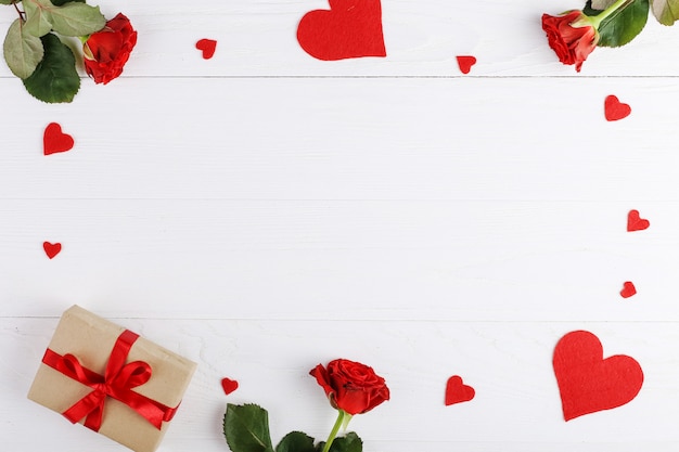 A bouquet of red roses, a gift and hearts on a white wooden table.