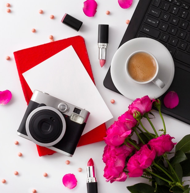 Bouquet of roses, cup of coffee, laptop, camera, notepad and lipstick on white table.