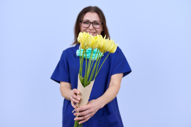 Bouquet of tulip flowers in hands of female doctor with removed protective medical mask World health day happy nurse with flowers on blue background