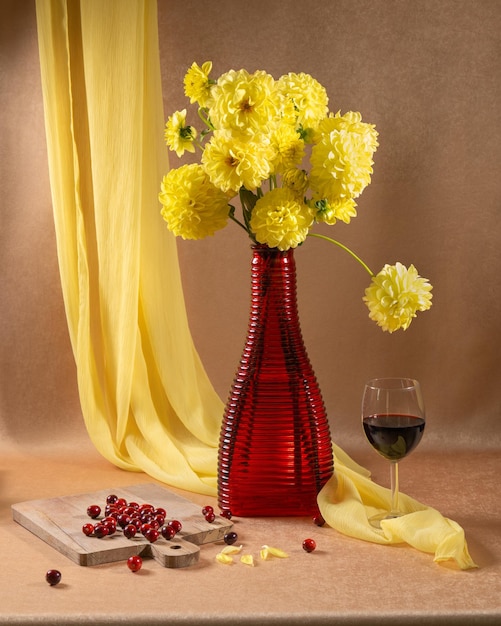 bouquet of yellow flowers in a red vase next to a glass of red wine and red berries on an orange bac