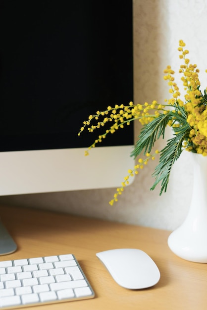 Bouquet of yellow mimosa flowers in a vase on a desktop with a computer