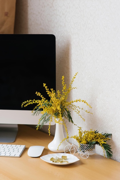 Bouquet of yellow mimosa flowers in a vase on a desktop with a computer