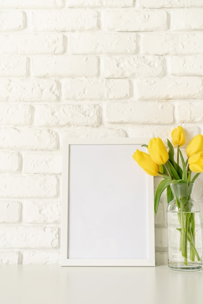 Bouquet of yellow tulips in a glass vase and blank photo frame on a white brick wall background. Mock up design