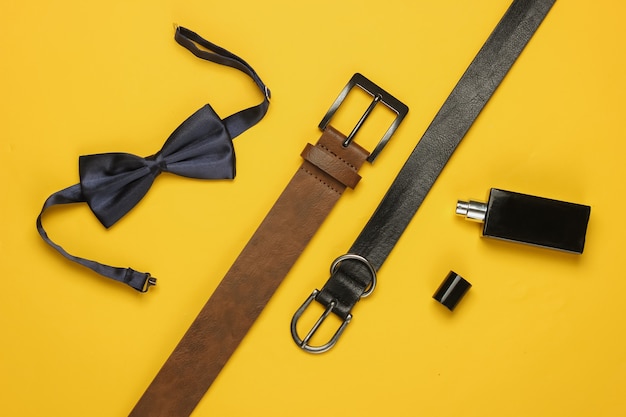 Bow tie, belts, perfume bottle on yellow background. Gents accessories, men's business style set. Formal style, preparing for the wedding.