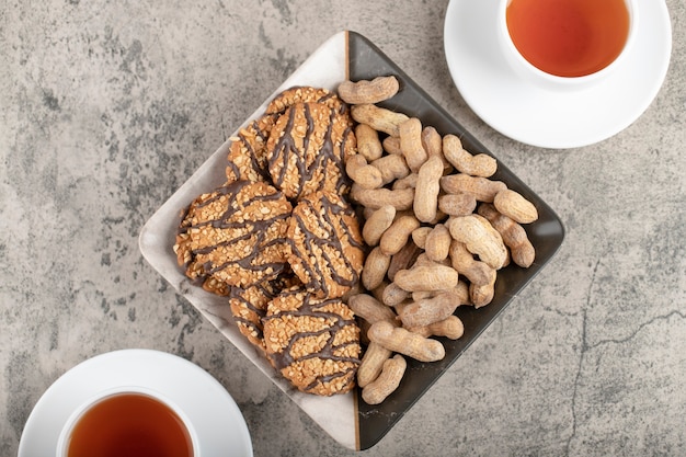 Bowl of biscuits and peanuts with cup of tea on stone surface.