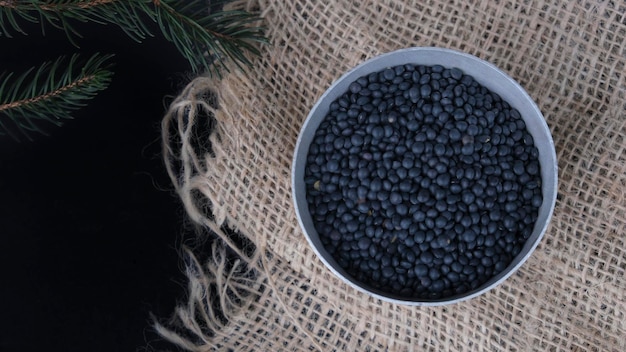 Bowl of black lentils on a rustic cloth sack on a black background with pine branches with copy spac