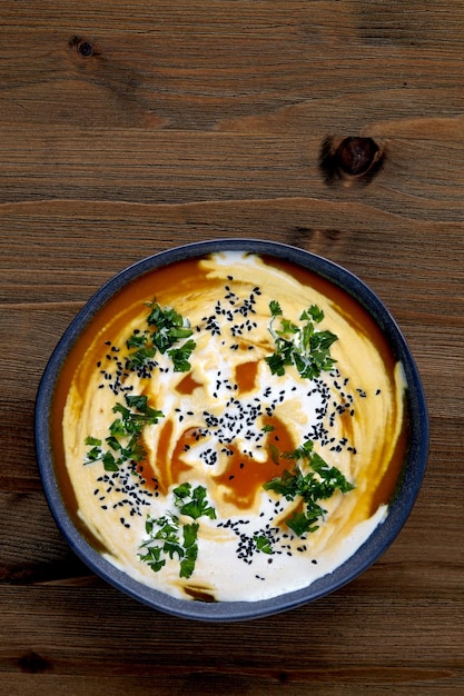 Bowl of creamy pumpkin soup with cream black cumin seeds herbs and cranberries on a wooden table