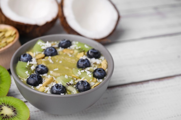 Bowl of delicious fruit smoothie with fresh blueberries kiwi slices and coconut flakes on white wooden table closeup Space for text