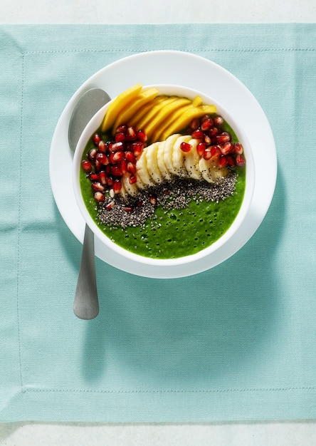 Bowl of green smoothie with sliced mango, banana, pomegranate seeds and chia seeds, maple syrup for a healthy morning breakfast