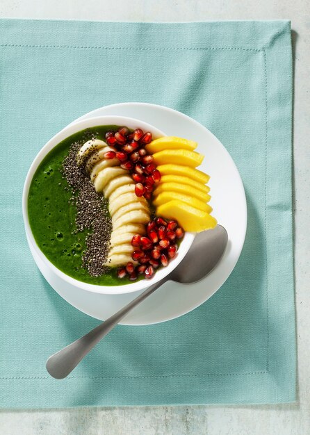 Bowl of green smoothie with sliced mango, banana, pomegranate seeds and chia seeds, maple syrup for a healthy morning breakfast