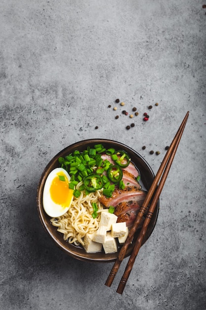 Bowl of tasty Asian noodle soup ramen with broth, tofu, pork, egg on grey rustic concrete background, space for text, close up, top view. Hot tasty Japanese ramen soup for dinner with copy space