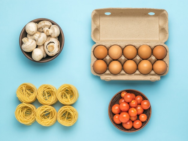 A bowl of tomatoes, a bowl of mushrooms, pasta and a box of eggs on a blue background. Ingredients for making pasta.