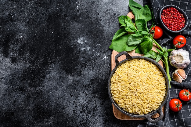 A bowl of uncooked Orzo pasta with Basil leaves, tomatoes and garlic. Black background. Top view. Copy space