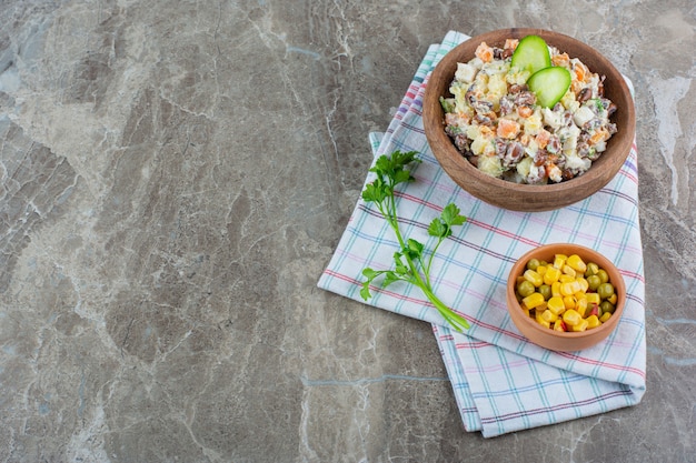 A bowl of vegetable salad next to a corn salad in a bowl on a tea towel , on the marble background.