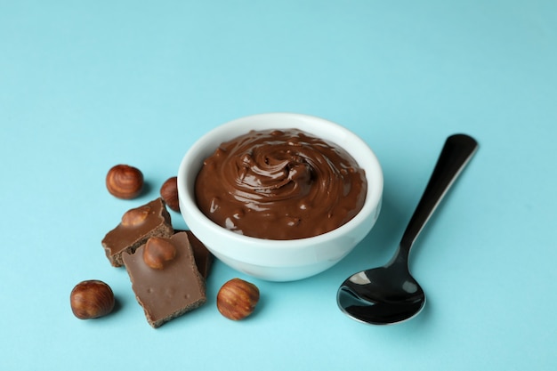 Bowl with chocolate paste, nuts, chocolate and spoon on blue background