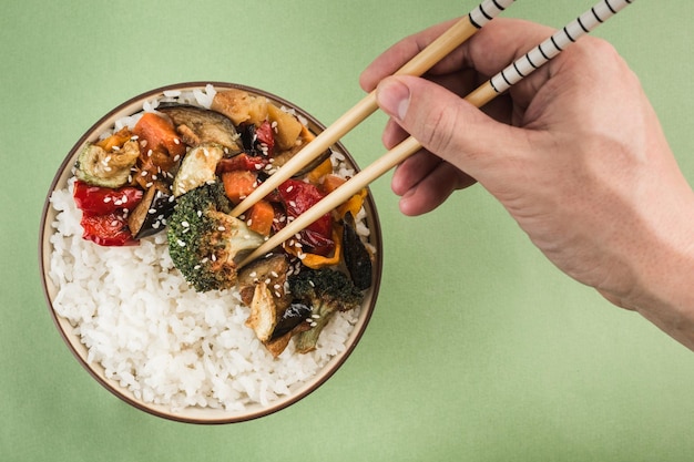 A bowl with grilled rice and vegetables on a light green background. A hand with chopsticks takes broccoli from a plate. Asian food. Horizontal orientation. Top view with a copy space for the text.