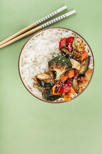 A bowl with grilled rice and vegetables with chopsticks on a light green background. Asian food. Vertical orientation. Top view with a copy space for the text.