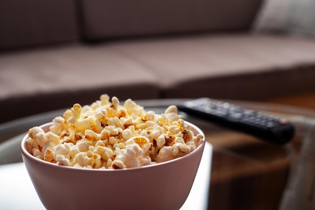 Bowl with popcorn and tv remote control on glass table near sofa