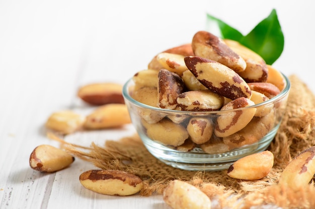 Bowl with tasty Brazil nuts on sack and space for text on white wooden background Healthy food