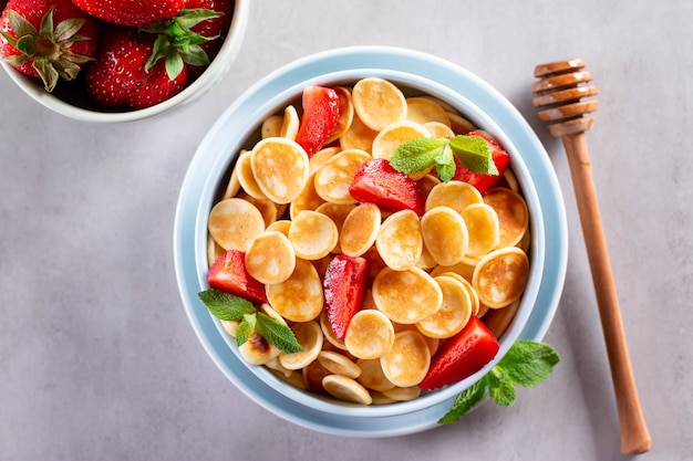 Bowl with tiny pancake cereal on a light concrete background. Trendy food. Mini cereal pancakes with honey and strawberries. Top view