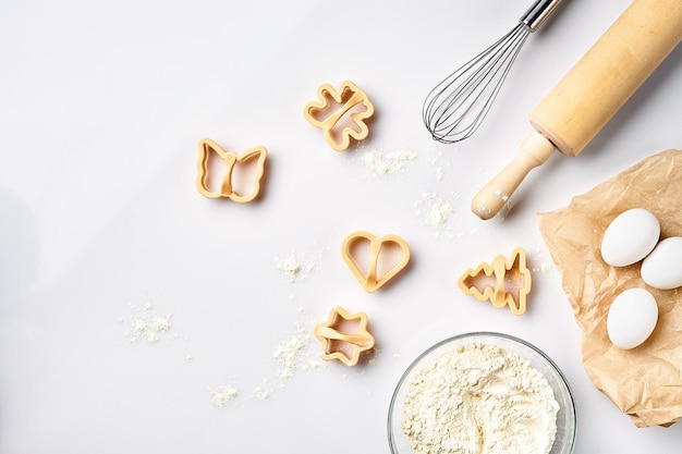 Bowl with wheat flour rolling pin whisk eggs cookie cutters Top view on a white table with a copy space