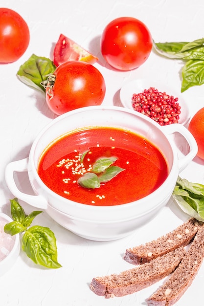 Bowls with homemade tomato soup. Ripe vegetables, fresh basil leaves, breadsticks, aromatic spices. Trendy hard light, dark shadow. White putty background, flat lay