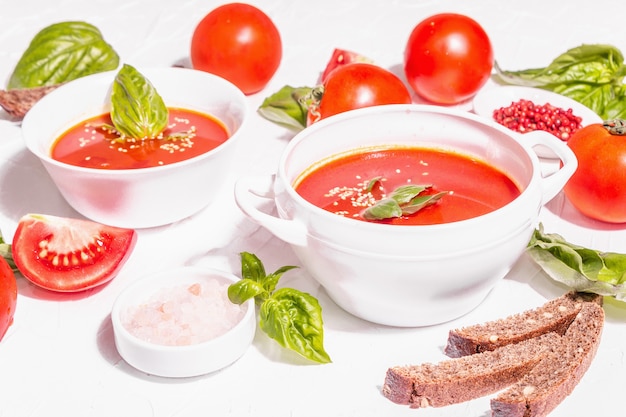 Bowls with homemade tomato soup. Ripe vegetables, fresh basil leaves, breadsticks, aromatic spices. Trendy hard light, dark shadow. White putty background, flat lay