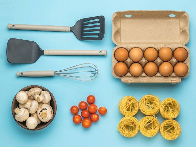 A box of eggs, a bowl of mushrooms, pasta, tomatoes, and cooking utensils. Ingredients for making pasta.