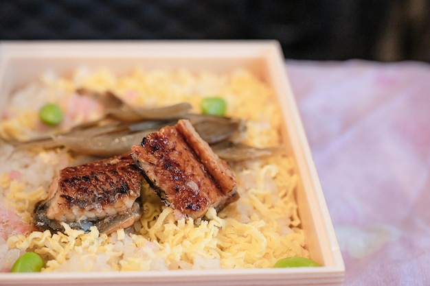 A box of rice with a piece of meat and a green pea on top.