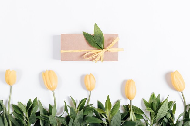 Box with a gift and a row of yellow tulips on a white background, top view