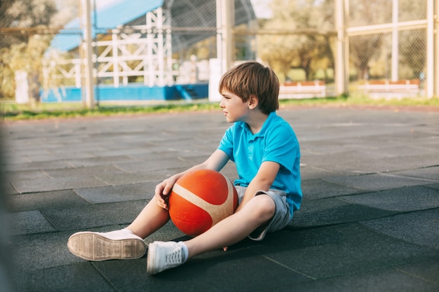 A boy basketball player in a blue t-shirt is sitting on the basketball court with a ball