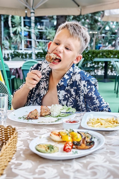 Boy bites and chews piece of meat on fork eating at outdoor table in restaurant