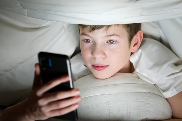 Boy under the blanket at night in his bed communicates on internet child gadget addiction and insomn...