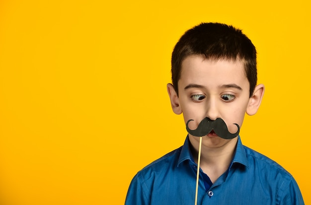 A boy in a blue shirt on a yellow background is twisted and dresses his mustache