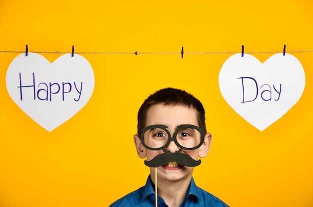 A boy in a blue shirt on a yellow background with a heart and an inscription a festive day is crooked and dresses glasses and a mustache