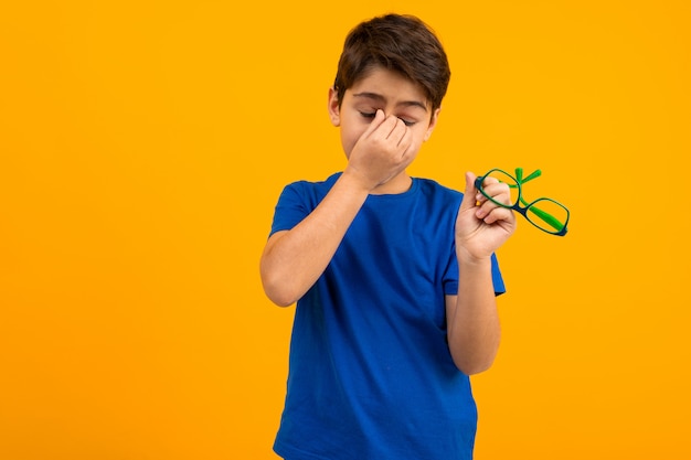 A boy in a blue T-shirt holds glasses in his hand and rubs his eyes on yellow with copy space