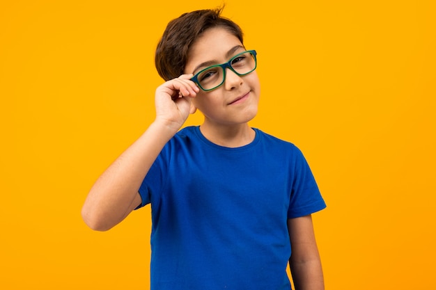 A boy in a blue t-shirt holds glasses with his hand and looks suspiciously at the camera on a bright yellow