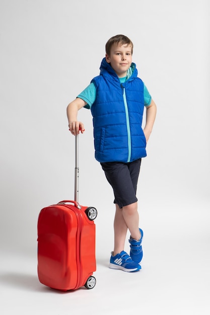 A boy in casual summer clothes shorts sneakers and a blue warm down vest with a red travel suitcase on wheels poses on a white background