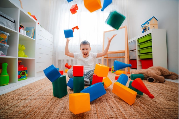 A boy child laughs sitting on the floor in the children's room throwing up toy colored plastic cubes