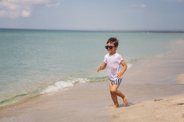 Boy child in striped shorts and a white T-shirt walks on sandy beach and in sunglasses