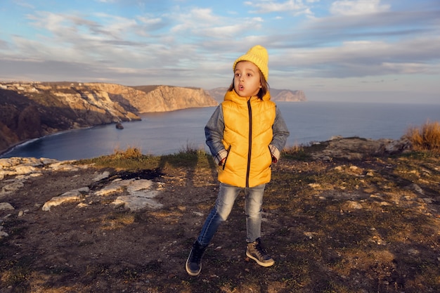 Boy child in a yellow jacket vest stands on a rock mountain by the sea in spring