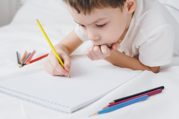 Boy draws with colorful pencils sitting on the bed 