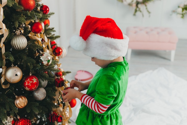 A boy dressed as a gnome and wearing a New Year's hat decorates a Christmas tree at home. Christmas.