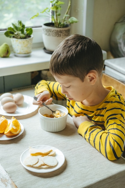 Photo boy eats breakfast in the morning with cereals with milk. morning breakfast in the kitchen before school. the boy is eating at the table by the window.