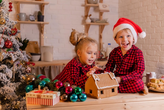 boy and girl in red christmas pajamas prepare a gingerbread house and decorate it with icing at home