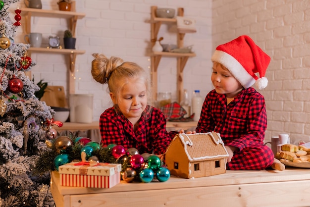 boy and girl in red christmas pajamas prepare a gingerbread house and decorate it with icing at home