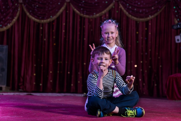 Boy and Girl Sitting Together on Stage, Girl is Making Rabbit Ears or Peace Signs Behind Boys Head