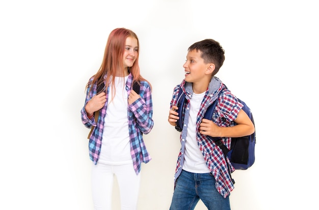 Boy and girl teenager 11 years old schoolboy and schoolgirl, looking on each other on white background with backpacks and smiling. Dressed in plaid shirt and white shirt