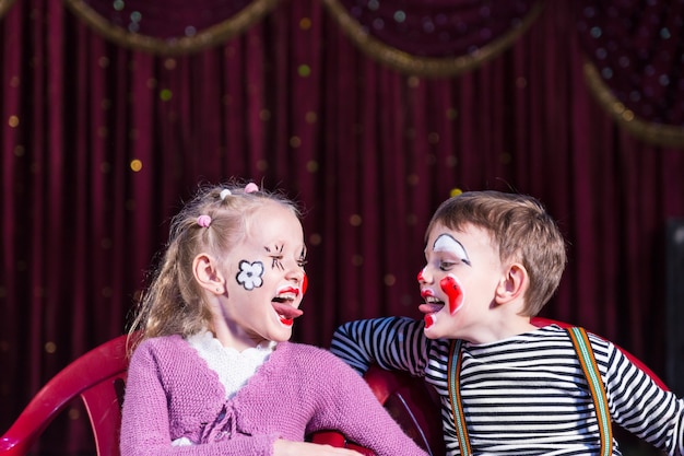 Boy and Girl Wearing Clown Make Up Sitting in Chairs Side by Side and Sticking Tongues Out at Each Other