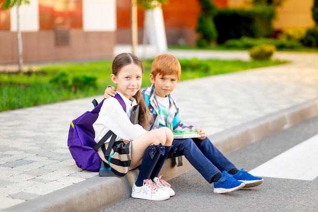 A boy and a girl with briefcases or backpacks are sitting at the school and reading a book and tablet going back to school brother and sister are studying doing homework on the street